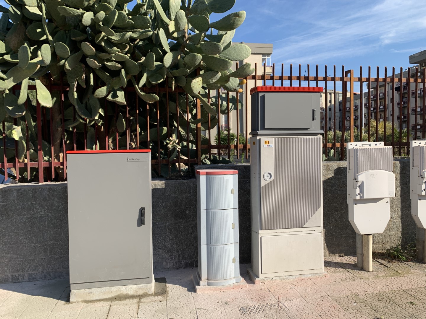 Marciapiede con installati diversi armadi stradali di TIM. Uno di questi è l'ARLO, è metallico, alto circa un metro, di colore grigio e con il tettuccio dipinto di rosso.